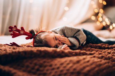 Küçük kız büyük bir yatakta yatıyor. Çocuğun boynuzları var. Noel havası.