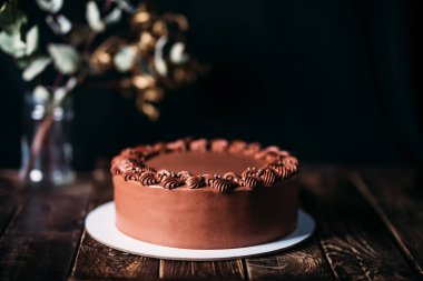 Beyaz tabakta çikolatalı pasta. Bulanık siyah bir arkaplanda, çiçekli bir vazo. Tahta masa ve güzel bir desen. Doğum günü ve diğer tatiller için geleneksel tatlılar.