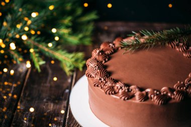 Beyaz tabakta çikolatalı pasta. Bir Noel ağacının arka planında çelenk ışıkları vardı. Tahta masa ve güzel bir desen. Doğum günü ve diğer tatiller için geleneksel tatlılar.