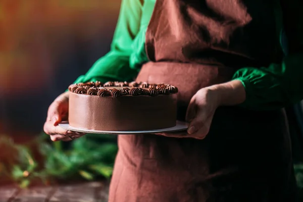 Woman baker in apron holding a chocolate cake on a dark background. Workers uniform in the kitchen. Against the background of a branch of a Christmas tree. Place for text.