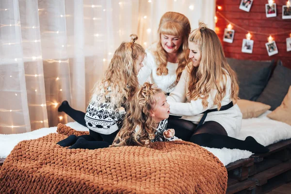 Children with mom and grandmother are sitting hugging on a large bed. Christmas mood. Against the background of a brick wall with garlands.