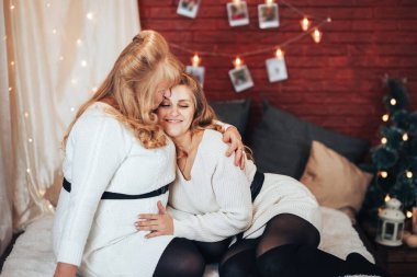 Anne ve kızı büyük yatakta oturuyorlar. Noel havası. Çelenkleri olan tuğla bir duvarın arkasında.