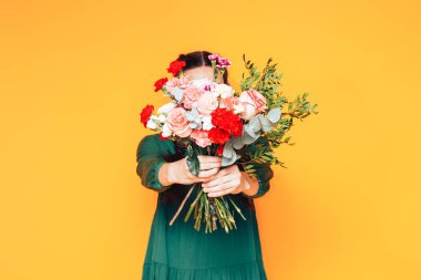 Sarı arka planda yeşil elbiseli büyük bir buket çiçek tutan bir kadın.