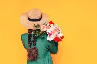Sarı arka planda yeşil elbiseli büyük bir buket çiçek tutan bir kadın..