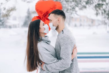 Parkta birbirine aşık genç bir çift kar ve kırmızı kalp şeklinde balonlardan oluşan bir arka planda. Sevgililer günü hediyesi. Tıbbi maskeli aşıklar öpüşür.