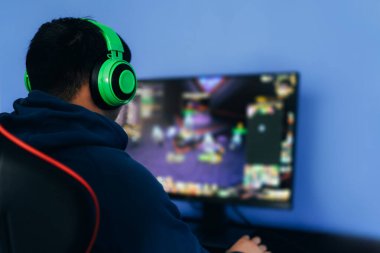 Profesyonel koltukta bilgisayar oyunu oynayan genç bir adam yeşil kulaklıklı monitöre karşı, canlı yayın, esportlar. mavi arkaplan