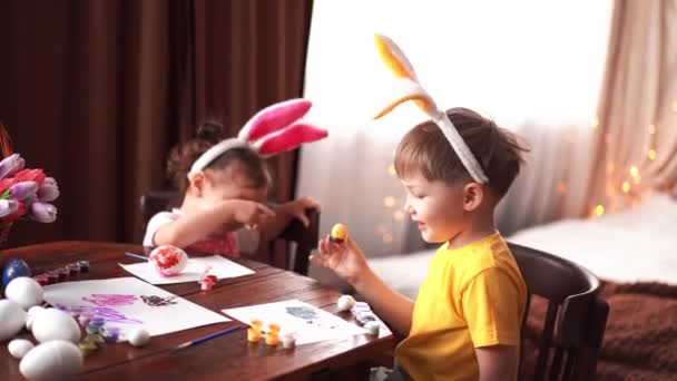 Brat i siostra malują pisanki na Wielkanoc. Szczęśliwe dzieci malują jajka w zabawny sposób. Dzieci mają królicze uszy na głowach. — Wideo stockowe