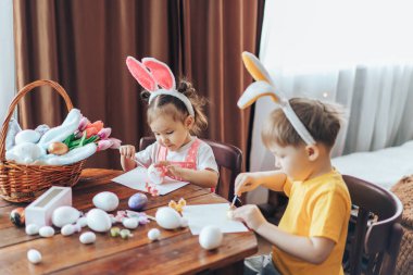 Paskalya için hazırlanan çocuklar dini bayram için yumurta boyuyor. Oğlan ve kız, bayram için tahta masada Paskalya yumurtası boyuyorlar. Çocukların kafalarında tavşan kulakları vardır..
