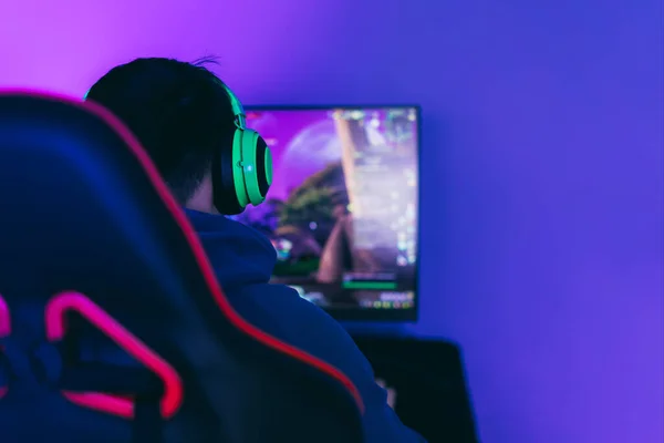 El jugador se sienta en una silla de juego y juega juegos de ordenador. El jugador tiene auriculares verdes en la cabeza —  Fotos de Stock