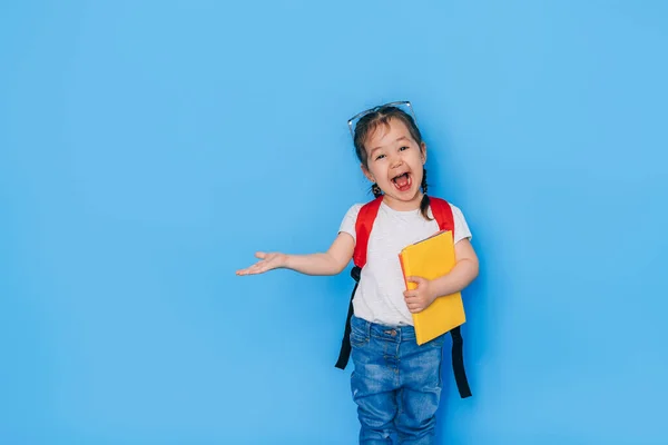 Surprised schoolgirl holding yellow book in one hand and pointing to the side against blue background with the other. Back to school in 2021. Homeschooling, distance learning.