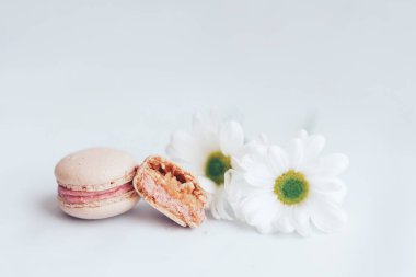 İki pembe badem kurabiyesi, biri bütün diğeri de ikiye bölünmüş. Kurabiyelerin yanında beyaz arka planda iki beyaz tomurcuk var..