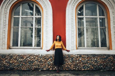 Sarı kazaklı ve kırmızı şapkalı bir kadın büyük pencereli bir binanın arka planında duruyor..