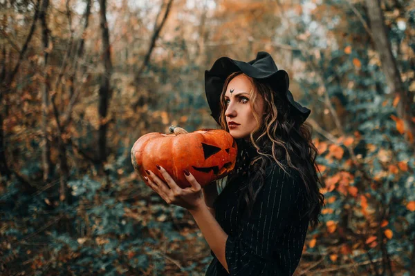Chica vestida como bruja sosteniendo calabaza con dos manos en el fondo del bosque —  Fotos de Stock