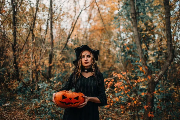 Chica vestida como bruja sosteniendo calabaza con dos manos en el fondo del bosque —  Fotos de Stock