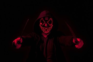 Man armed with two machetes and wearing a scary lighting neon glow mask and a hoodie on black background. clipart