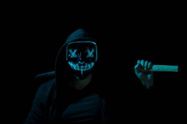 Man armed holding a katana and wearing a scary lighting neon glow mask and a hoodie on black background. clipart