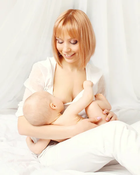 快乐的微笑母亲喂养母乳宝宝在家里在床上 — 图库照片