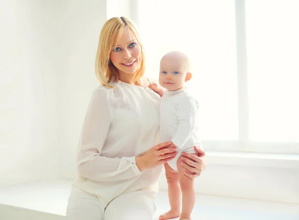 Feliz madre sonriente con bebé en casa en habitación blanca cerca de la ventana — Foto de Stock