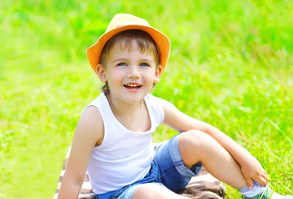 Glad liten pojke barn i hatt sitter på gräset i sommardag — Stockfoto