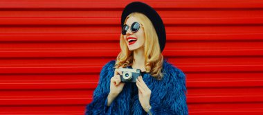 Retro kameralı, mavi kürklü, kırmızı arka planda siyah yuvarlak şapkalı, şaşırmış genç bir kadın.