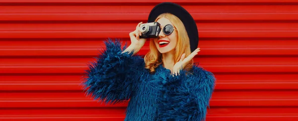 レトロカメラを身に着けている写真を撮ると驚きの女性の肖像ブルーフェイクファーコート 赤い背景の上にラウンド帽子 — ストック写真