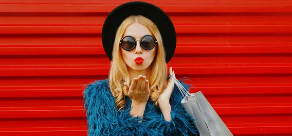 魅力的な女性モデルの肖像画でショッピングバッグ吹いて赤い唇甘い空気キス身に着けている青フェイクファーコート ラウンド帽子以上赤壁の背景 — ストック写真