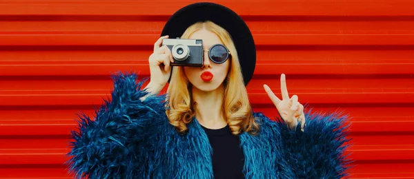 赤い唇を吹いて写真を撮るレトロなカメラを持つ女性のファッションポートレート送信甘い空気キス身に着けている青いフェイクファーコート上の赤い背景 — ストック写真