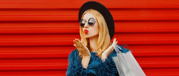 魅力的な女性モデルの肖像画でショッピングバッグ吹いて赤い唇甘い空気キス身に着けている青フェイクファーコート ラウンド帽子以上赤壁の背景 — ストック写真