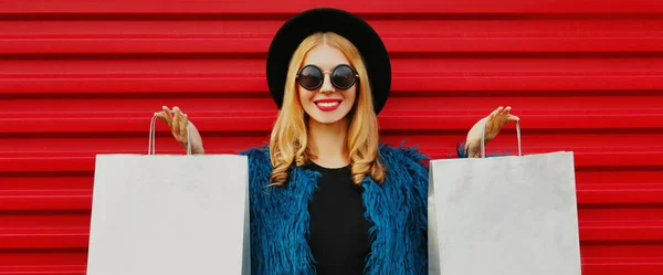 ポートレートクローズアップのスタイリッシュなブロンド女性とともに二つのショッピングバッグ身に着けています青フェイクファーコート 黒丸帽子以上赤壁の背景 — ストック写真