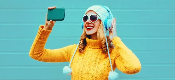 黄色のニットセーター 白い帽子 ハート型のサングラスを身に着けている無線ヘッドフォンで音楽を聞きながら 幸せな笑顔の女性は電話で写真を撮る青の背景に — ストック写真