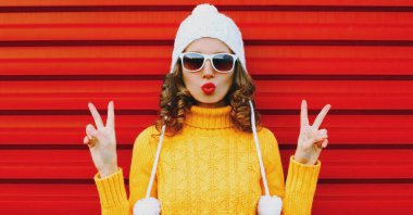 Kış portresi genç bir kadın kırmızı dudaklar üflüyor sarı örülü süveter ve kırmızı arka planda ponpon şapkası olan beyaz bir öpücük yolluyor.