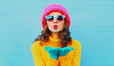 Kış portresi güzel bir kadın kırmızı dudaklar üflüyor sarı örülü süveter, mavi arka planda pembe şapka ile tatlı bir öpücük yolluyor.