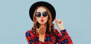 Genç bir kadının portresi kırmızı dudaklar üflüyor. Mavi arka planda siyah yuvarlak şapkalı tatlı bir öpücük yolluyor.