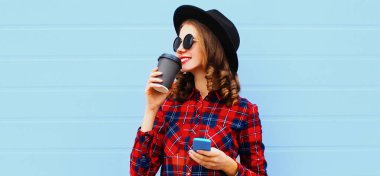 Mavi arka planda siyah yuvarlak şapkalı, telefonla kahve içen genç bir kadının portresi.