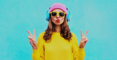 Kablosuz kulaklıkla müzik dinleyen, sarı örgü süveter ve mavi arka planda pembe şapka giyen modern genç bir kadının portresi.