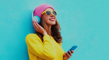Kablosuz kulaklıkla müzik dinleyen, sarı örgü süveter giyen, mavi arka planda pembe şapkalı modern genç kadının portresi.
