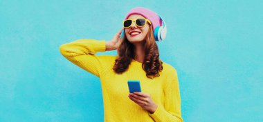 Kablosuz kulaklıkla müzik dinleyen, sarı örgü süveter giyen, mavi arka planda pembe şapkalı modern genç kadının portresi.