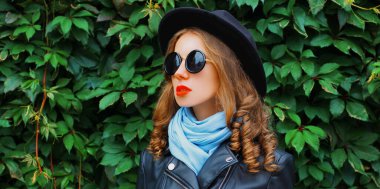 Arka plan olarak siyah yuvarlak bir şapka, ceket, yeşil yaprakların üzerine güneş gözlüğü takan güzel bir kadın modeli portresi.