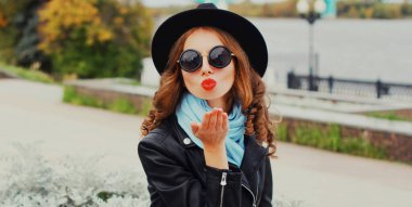 Güzel genç bir kadının portresi, dudaklarını silip, hava öpücüğü yolluyor. Siyah bir şapka, ceket ve güneş gözlüğü takıyor.