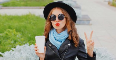 Kahve fincanı olan, siyah yuvarlak şapkalı, ceketli, güneş gözlüklü güzel bir kadının portresi.
