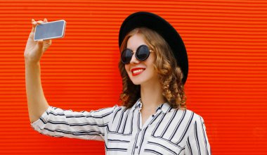 Mutlu gülümseyen kadının portresi telefonla fotoğraf çekiyor beyaz çizgili gömlekli, kırmızı arka planda siyah yuvarlak şapkalı.