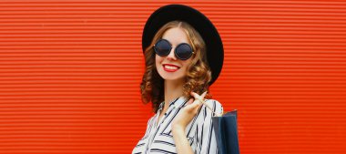 Kırmızı arka planda siyah yuvarlak bir şapka takan güzel, gülümseyen genç bir kadının portresi.