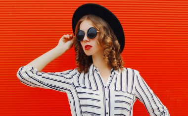 Siyah yuvarlak şapkalı ve kırmızı arka planda çizgili beyaz gömlekli çekici bir kadın portresi.