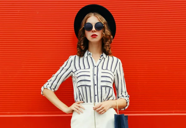 白い縞模様のシャツを着た買い物袋を持つ美しい若い女性の肖像画 赤い背景の上に街の通りに黒い丸帽子 — ストック写真