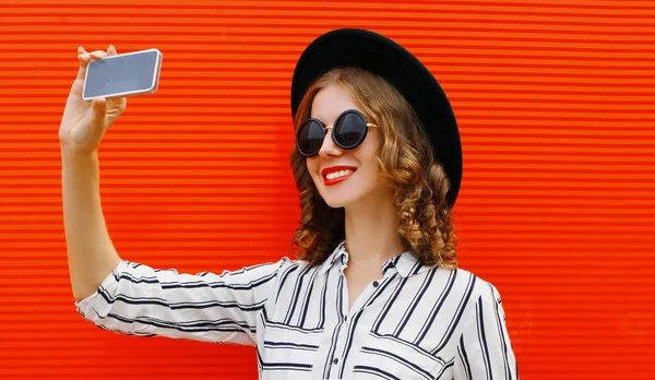 Mutlu gülümseyen kadının portresi telefonla fotoğraf çekiyor beyaz çizgili gömlekli, kırmızı arka planda siyah yuvarlak şapkalı.