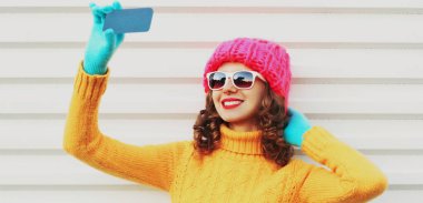 Kışın gülümseyen genç bir kadının portresi telefonla fotoğraf çekiyor. Renkli pembe bir şapka takıyor, beyaz arka planda sarı süveter örüyor.