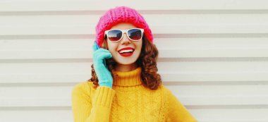 Mutlu gülümseyen genç bir kadının portresi. Akıllı telefondan arıyor. Renkli pembe bir şapka takıyor, beyaz arka planda sarı süveter giyiyor.