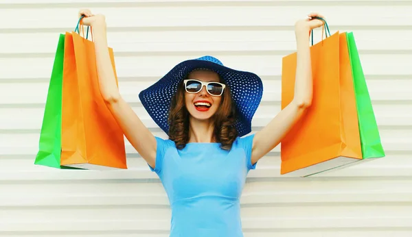 陽気若い笑顔の女性は白い背景に青いドレス 夏のわらラウンド帽子を身に着けている2つのショッピングバッグを示しています — ストック写真
