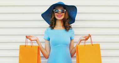 Çekici, gülümseyen bir kadın, iki tane turuncu alışveriş torbası, mavi bir elbise giyiyor, beyaz arka planda yaz hasır şapkası takıyor.