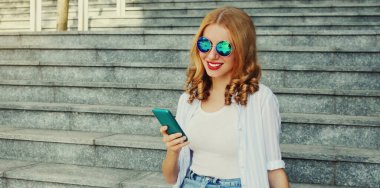 Şehirde merdivenlerde oturan akıllı telefonlu gülümseyen genç bir kadının portresi.